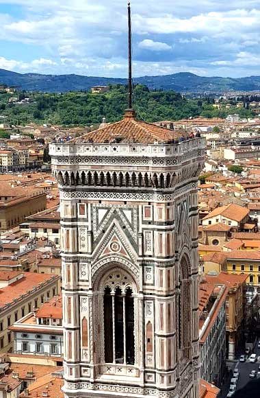 High-angle view of Giotto’s Campanile in Florence, Italy, showing its ornate white, green, and pink marble façade and terracotta roof, with the red-tiled city and green rolling hills under a partly cloudy sky