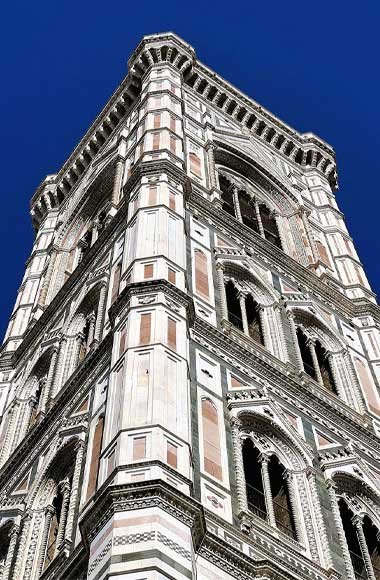 Low-angle view of Giotto’s Campanile in Florence, Italy, clad in white, green, and pink marble panels and intricate Gothic detailing against a deep blue sky