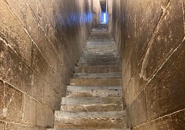 Interior of a historic bell tower showing a narrow, steep stone staircase with worn, uneven steps and rough masonry walls leading up toward a small, light-filled opening at the top