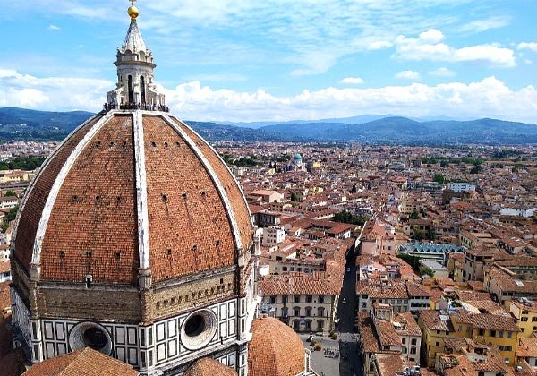 Aerial view of Florence’s Duomo, showing Brunelleschi’s red-tiled dome and lantern against a backdrop of the terracotta-roofed city and rolling Tuscan hills under a bright blue sky dotted with clouds