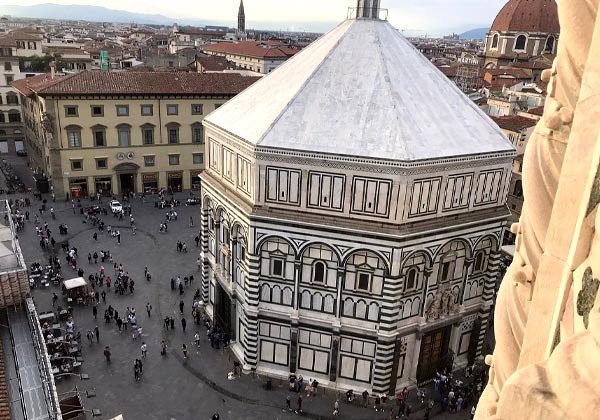 High-angle view of Florence’s octagonal Baptistery of San Giovanni clad in white and green marble panels, surrounded by visitors in the Piazza del Duomo and red-tiled city buildings beyond