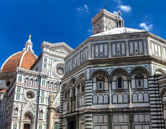 View of Florence’s Baptistery of San Giovanni with its green-and-white marble façade in the foreground, and the ornate Gothic façade and red-tiled dome of Santa Maria del Fiore rising behind it against a clear blue sky