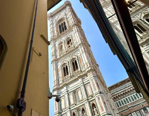 Upward view of Giotto’s Bell Tower in Florence framed between narrow buildings, showcasing its detailed white, green, and pink marble design.